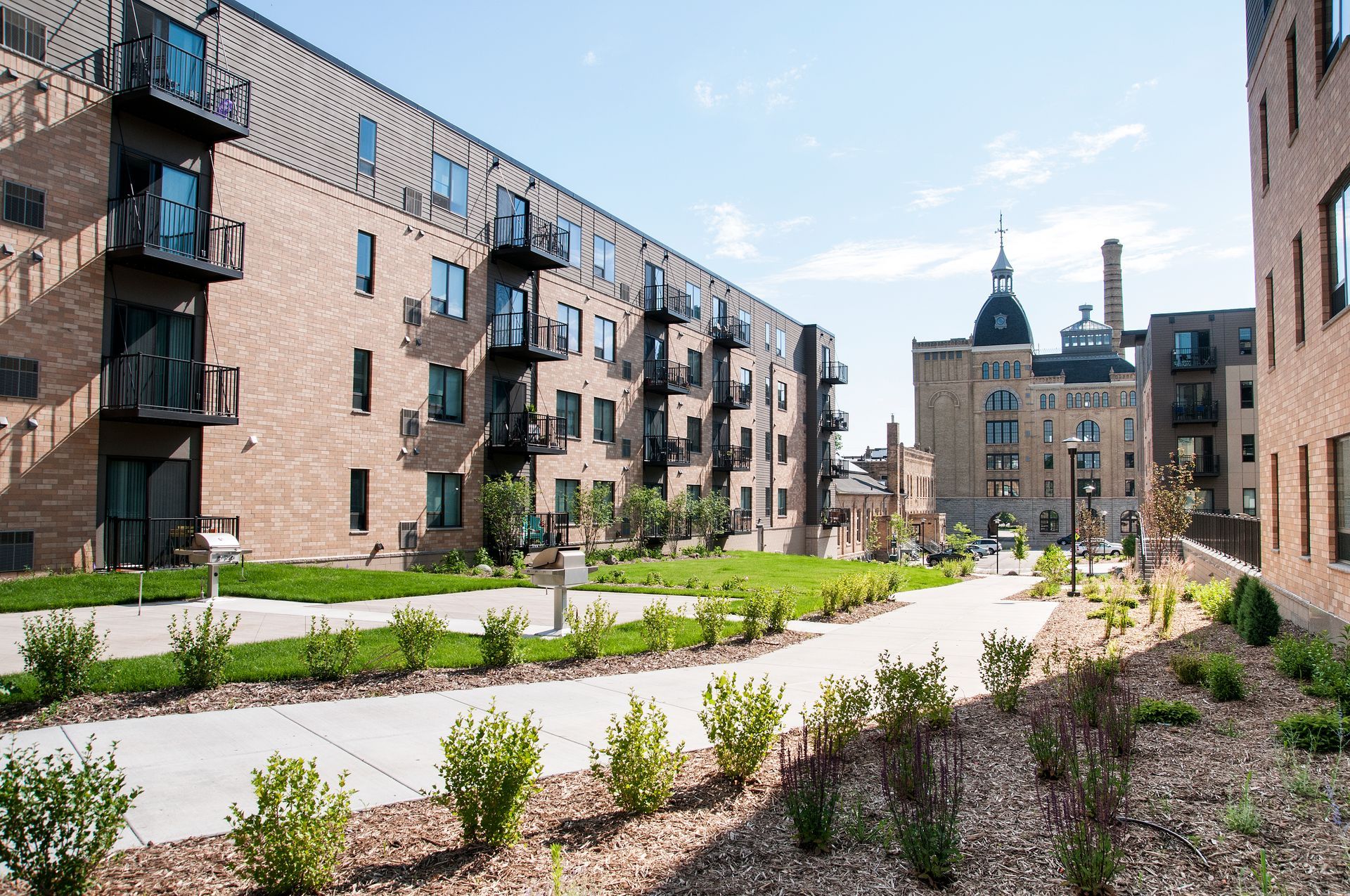 Apartment buildings with balconies and a green walkway leading to a domed building under a blue sky.