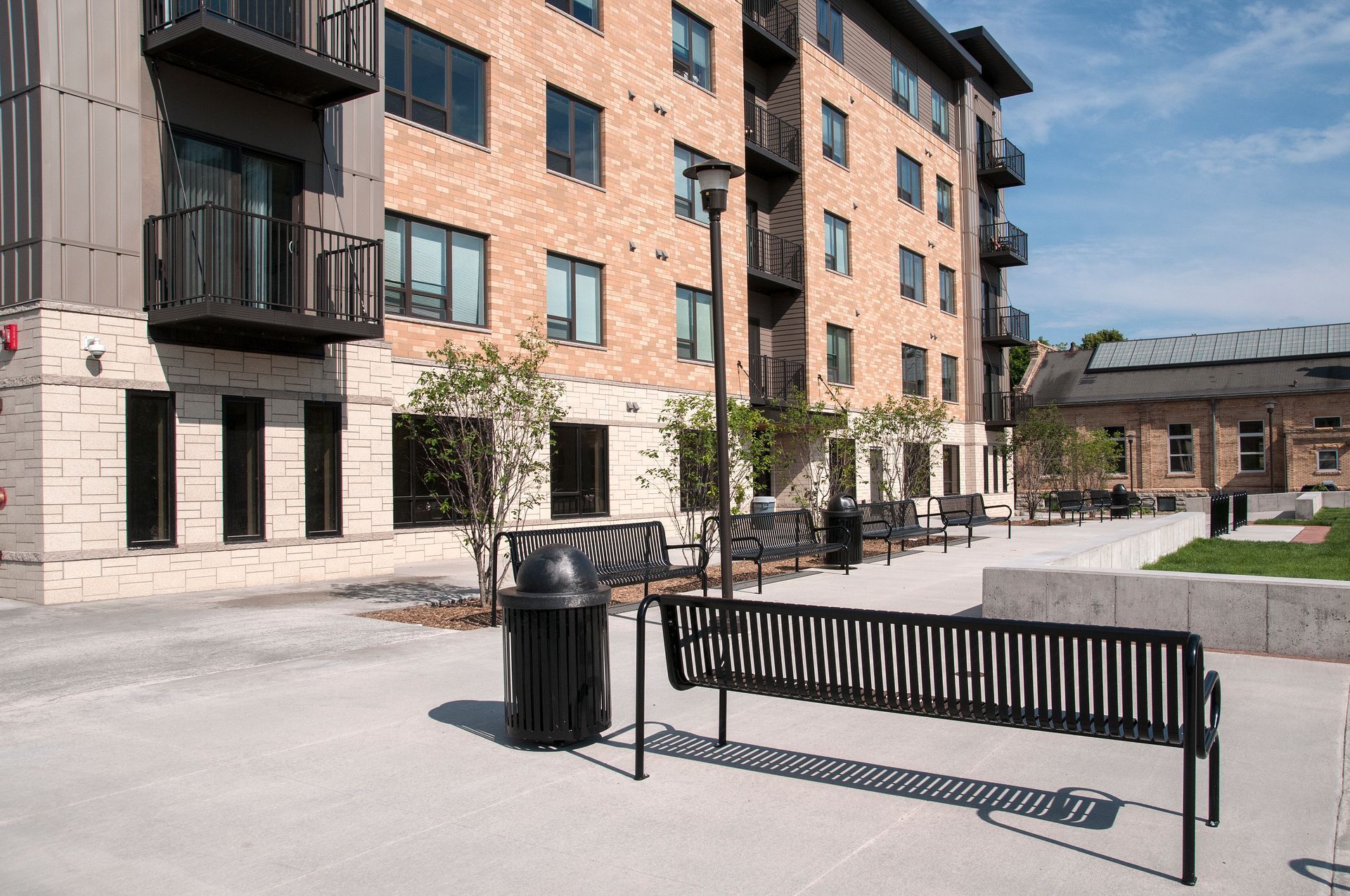 Exterior view of a modern apartment building with benches, landscaping, and a trash can.