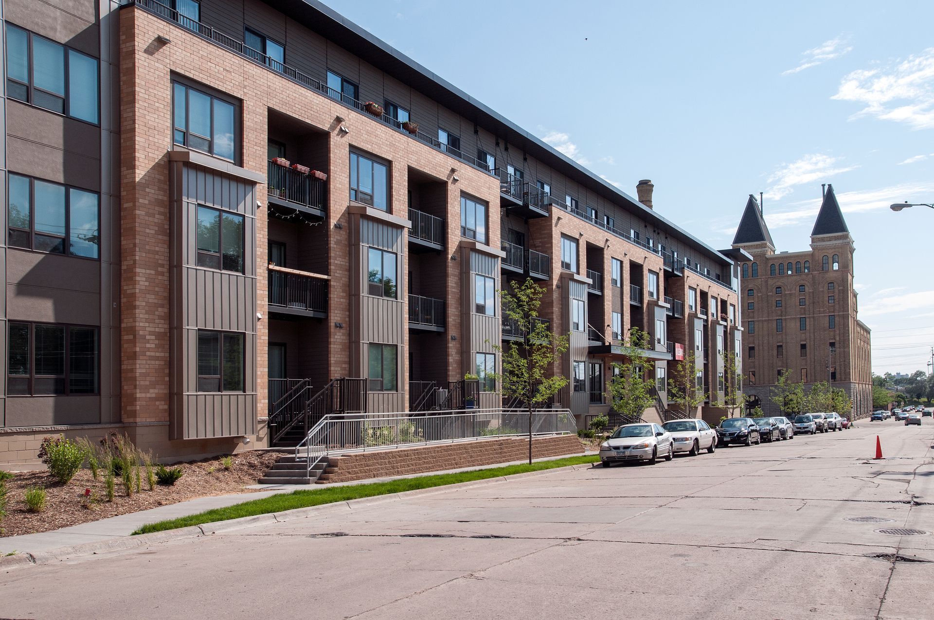 Modern brick apartment building and historic stone building along a street with parked cars.