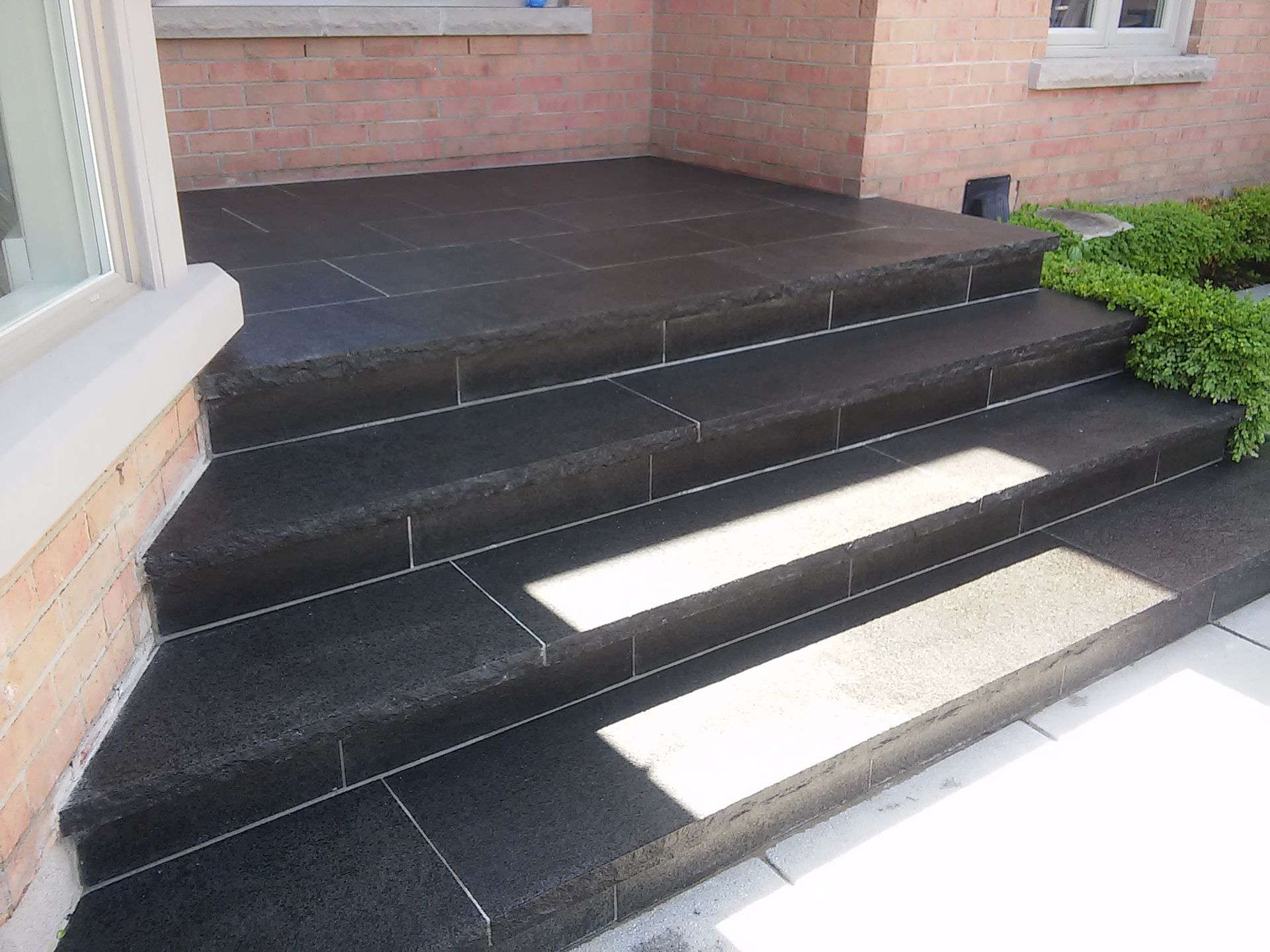 Black stone steps leading to a brick-walled porch, with bright sunlight and a green hedge.