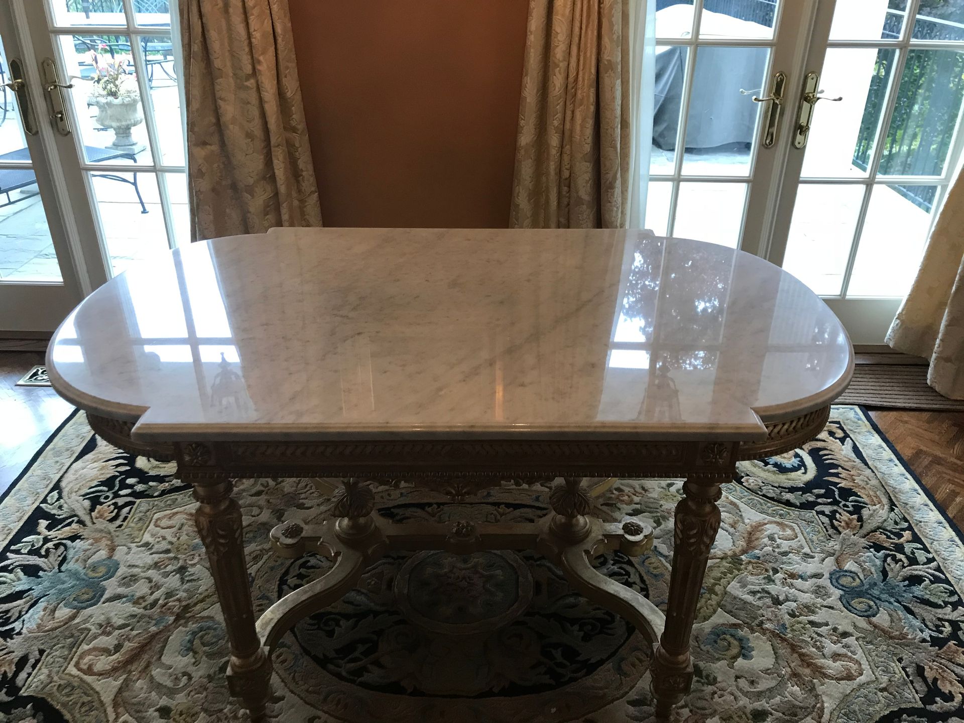 Marble-topped oval table with ornate wooden legs and a lower shelf, on a patterned rug near glass doors and curtains.