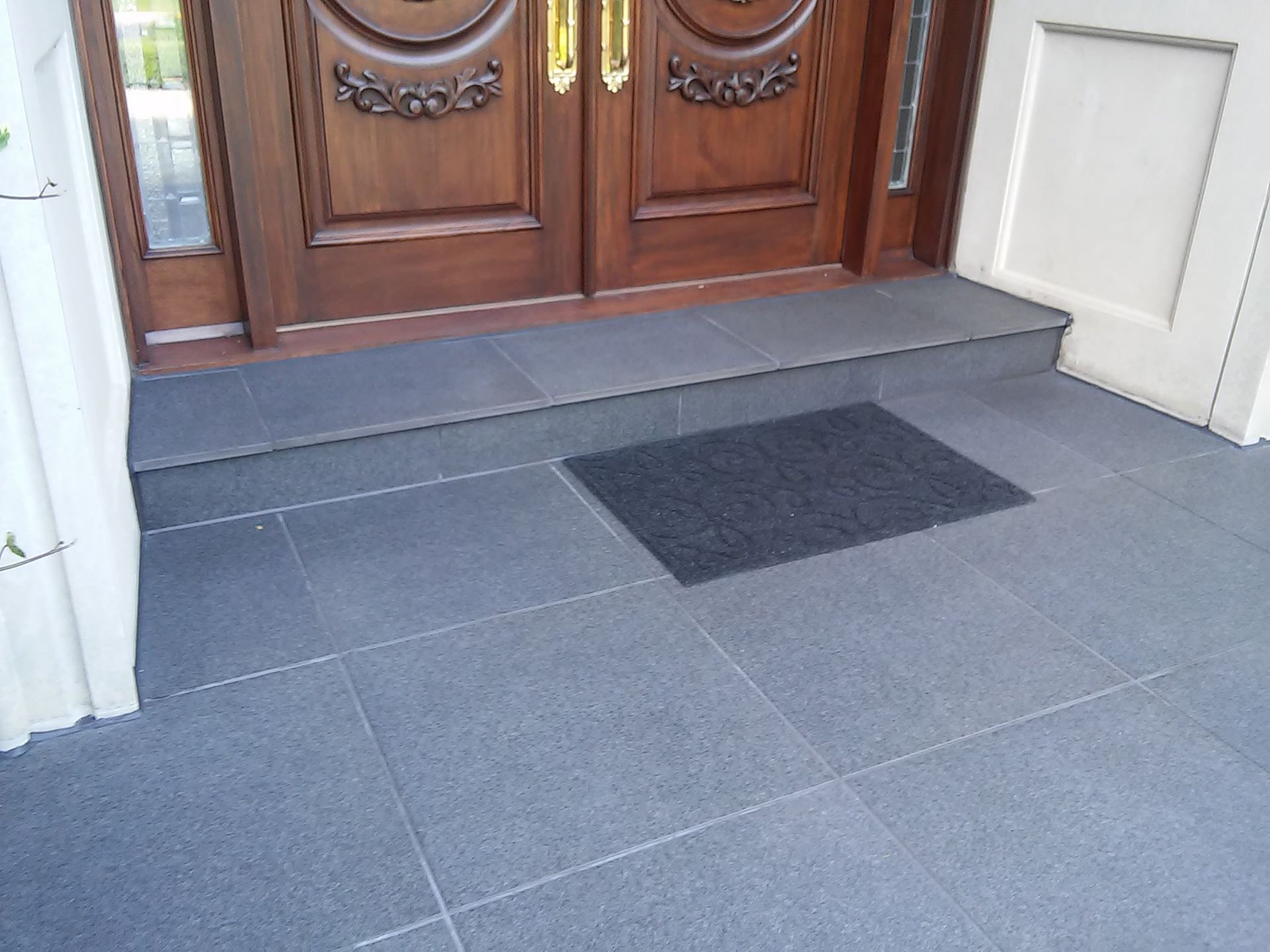 Dark tiled front entrance with a doormat in front of wooden double doors.
