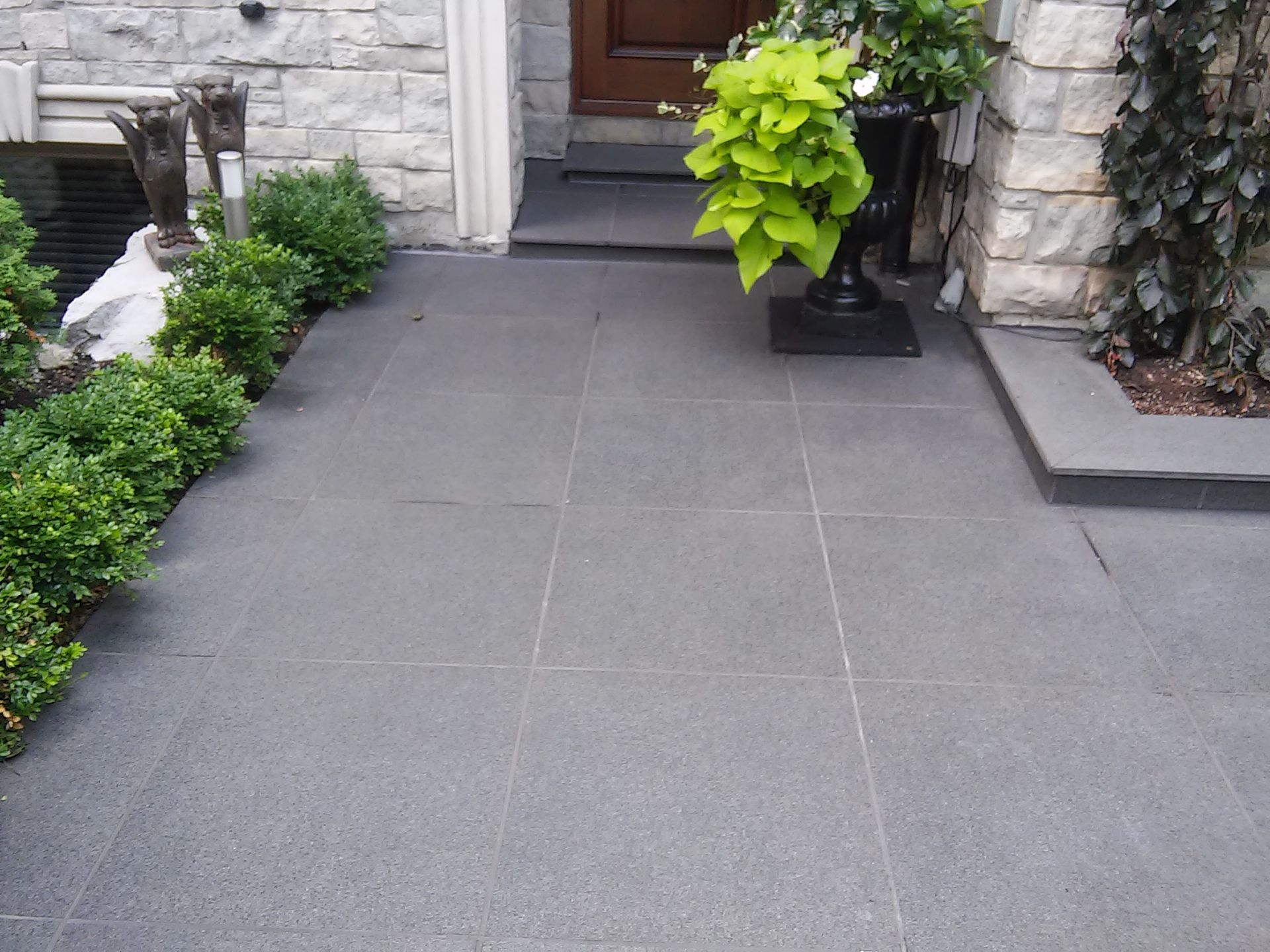 Gray tiled entryway with green bushes and potted plant, leading to a wooden door.