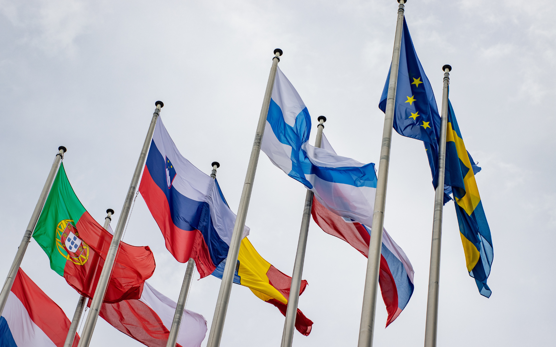 Flags of various European countries waving against a cloudy sky.