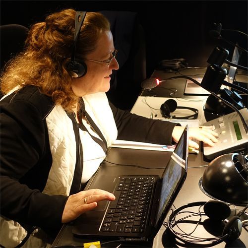 Woman wearing headphones at a desk, operating a laptop and other equipment in a studio setting.