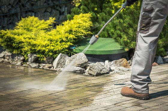 Person pressure washing a brick patio near shrubs and rocks.
