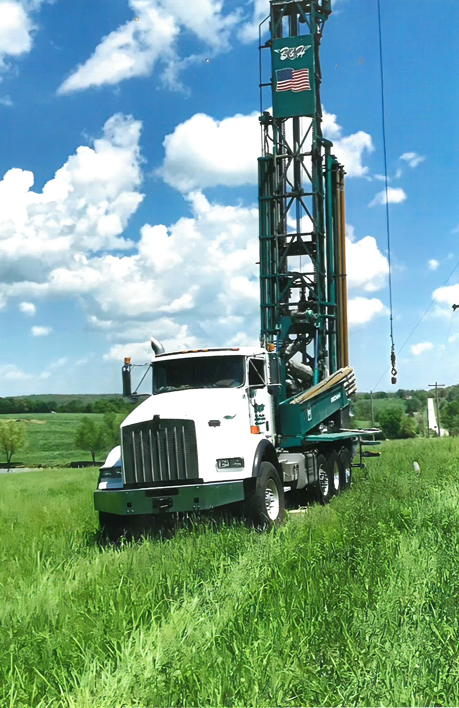 White truck with drilling rig on a grassy field under a blue sky with fluffy clouds.