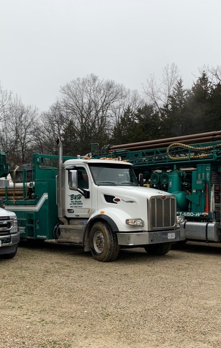 White truck with drilling rig on a grassy field under a blue sky with fluffy clouds.
