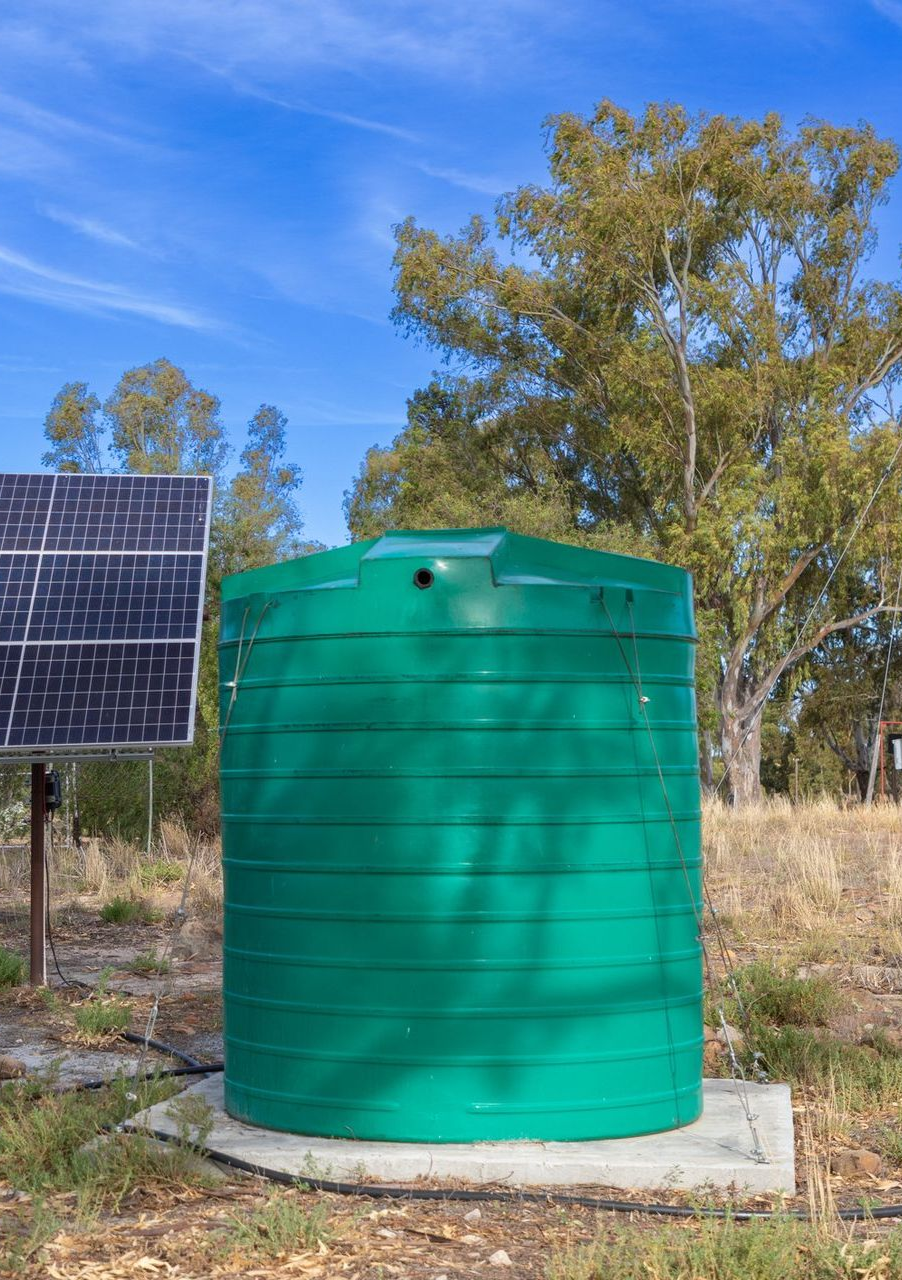 Green water tank, solar panel, and trees in a sunny outdoor setting.