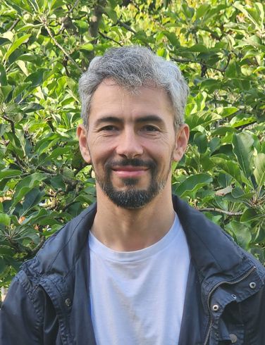 A man with a beard and gray hair is smiling in front of a tree.