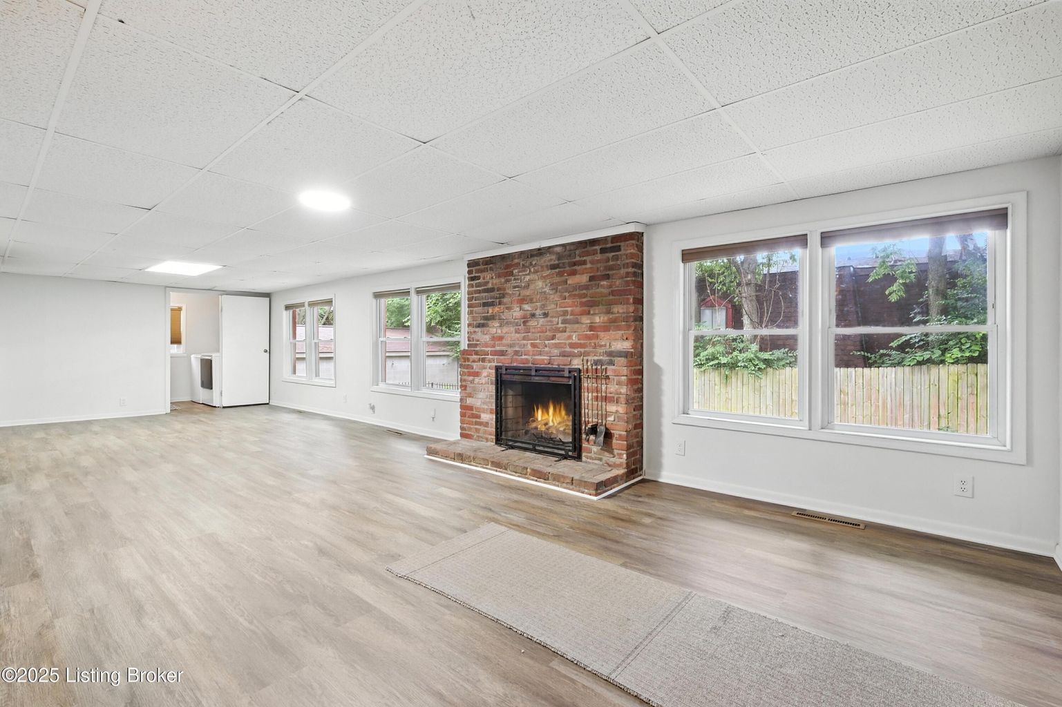 Spacious living room with brick fireplace, light gray flooring, and multiple windows overlooking greenery.