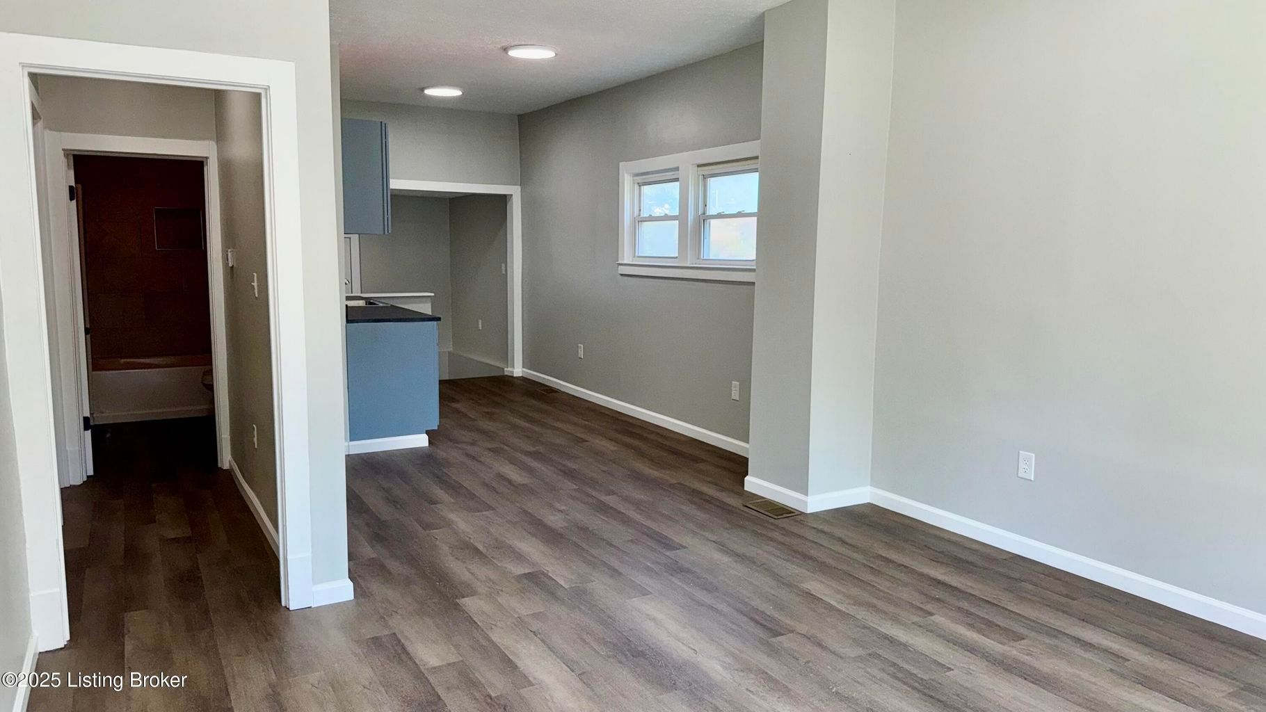 Empty room with wood-look flooring, light gray walls, a small kitchen, and a hallway to a bathroom.