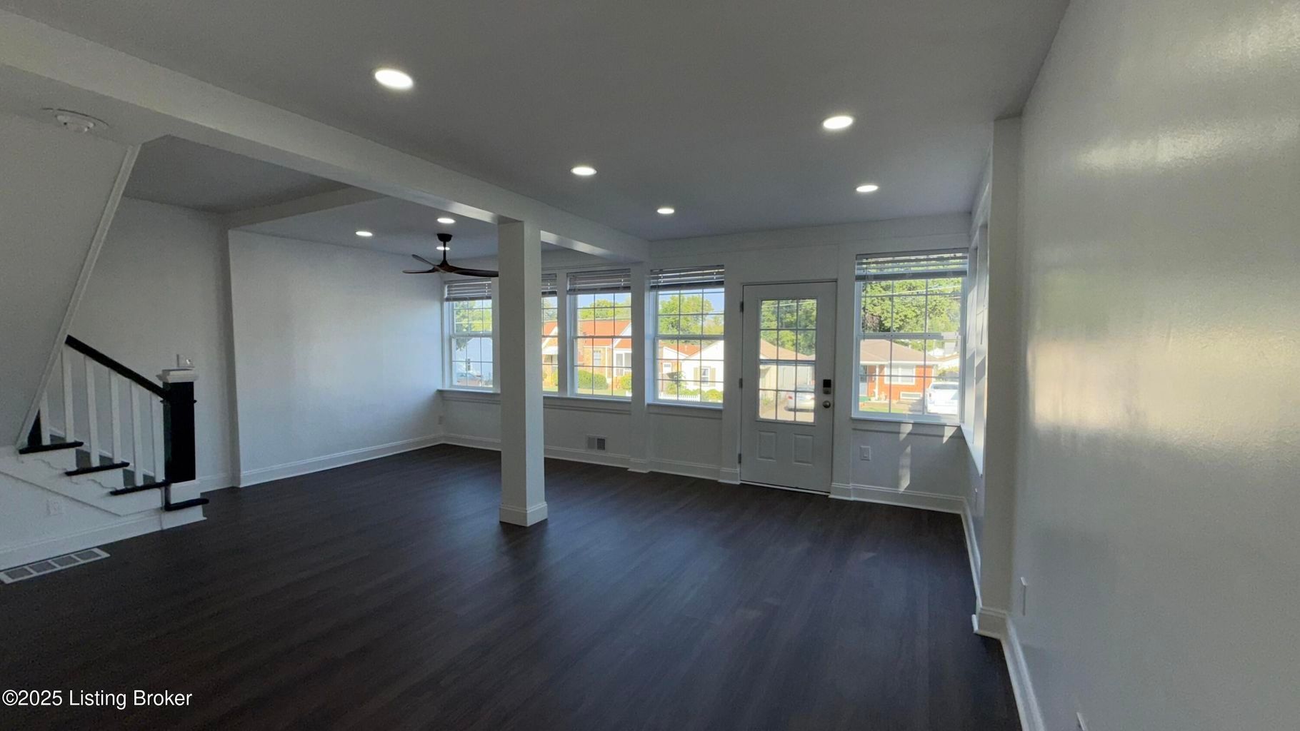 Empty renovated room with dark wood floors, white walls, and stairs. Windows and a door provide natural light.