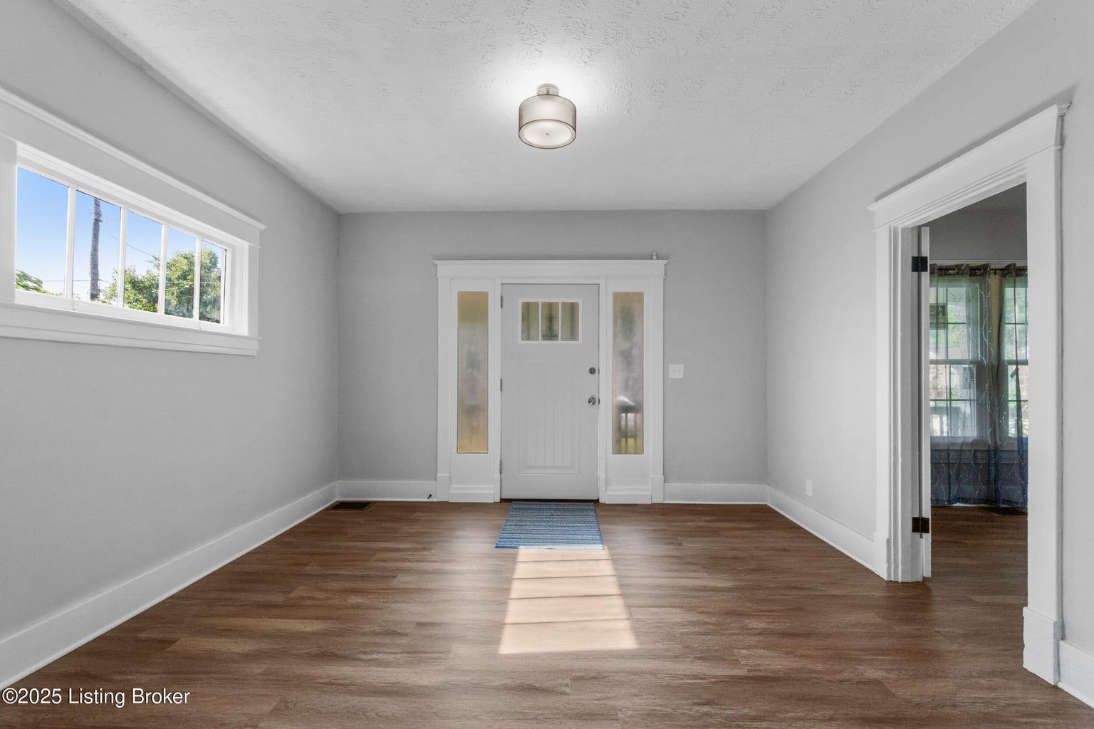 An interior view of a room with light gray walls, dark wood-tone floors, a white front door, and a white-framed window.