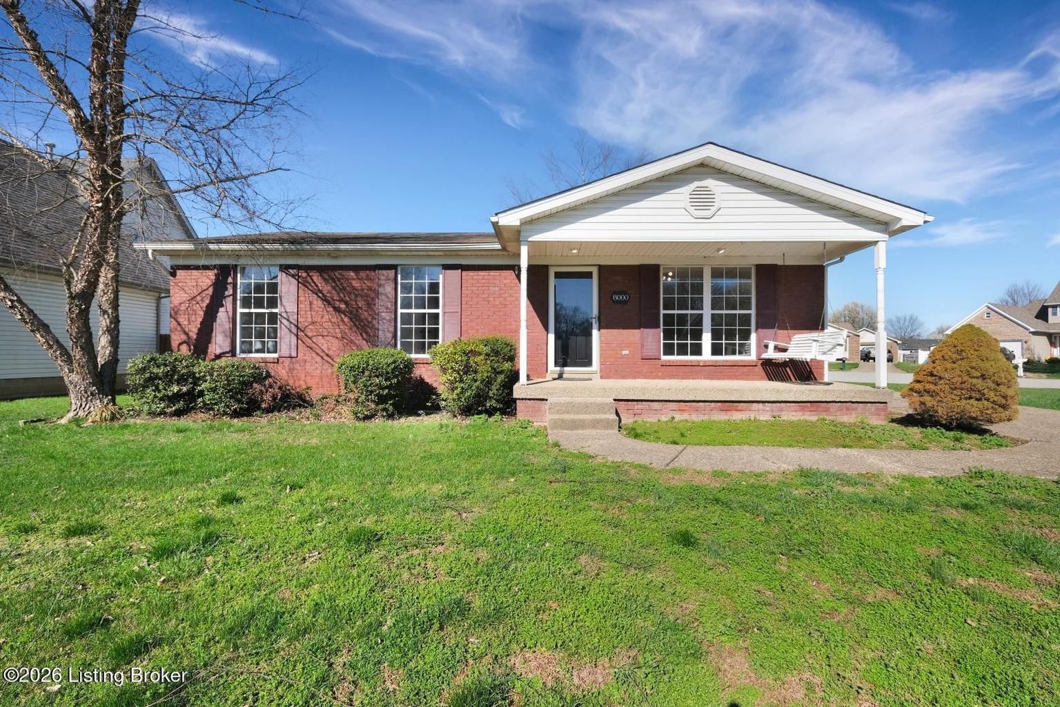 A single-story, red brick house with a white front porch and siding sits under a blue sky with thin, wispy clouds.