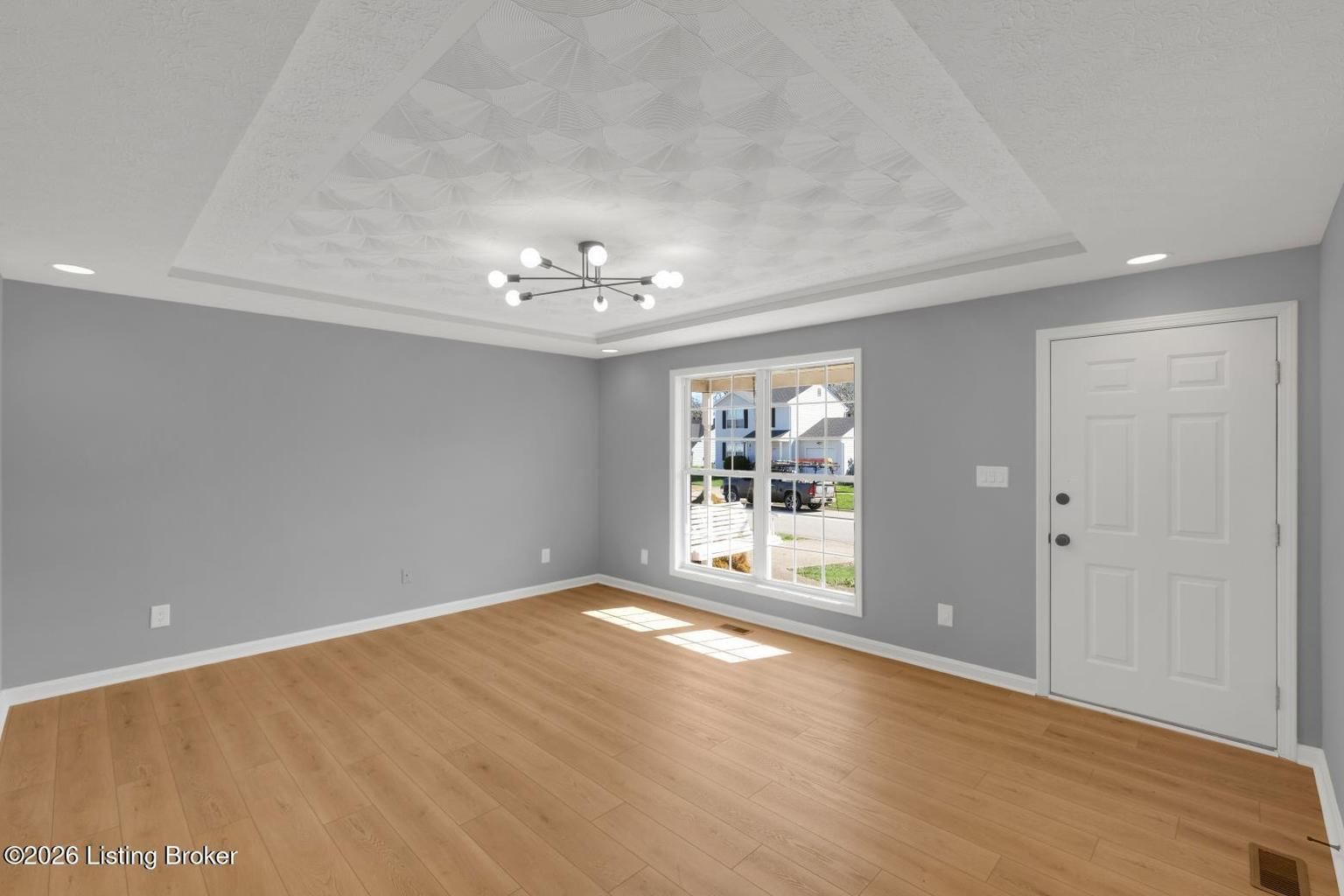 A bright, empty living room with gray walls, light wood flooring, a tray ceiling, modern light fixture, and front door.