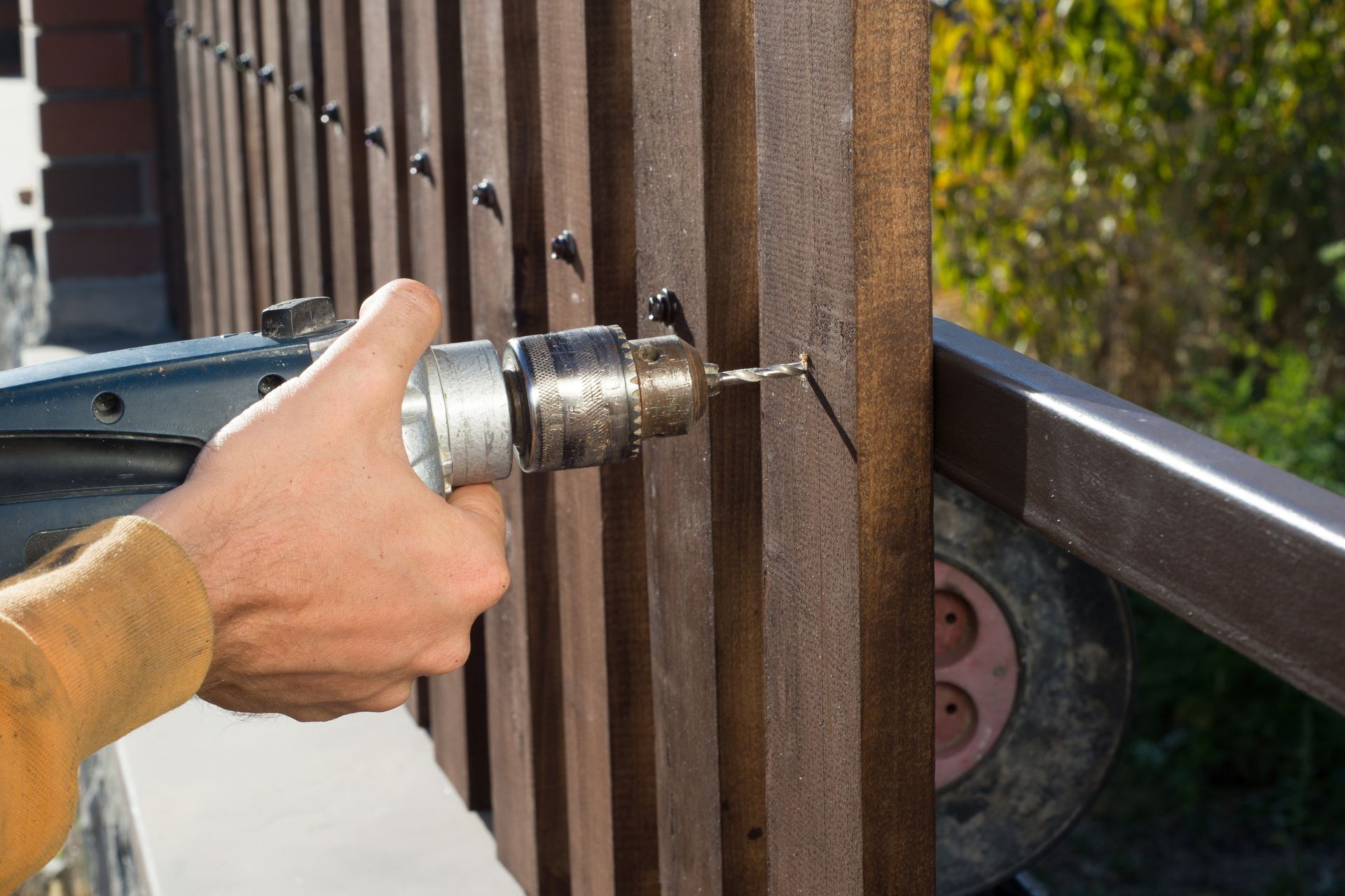 Hand using a power drill to bore into a rusty metal fence panel outdoors