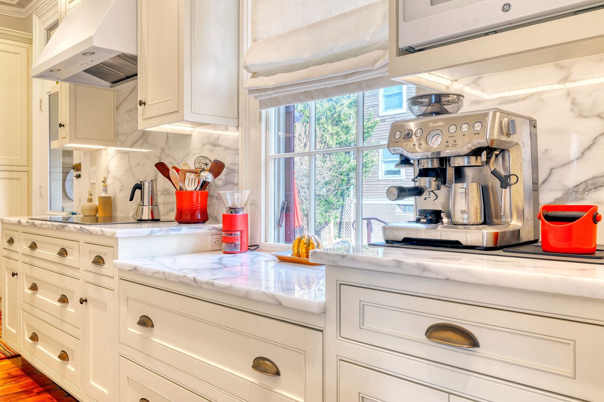 A kitchen with white cabinets , marble counter tops , a coffee maker and a window.