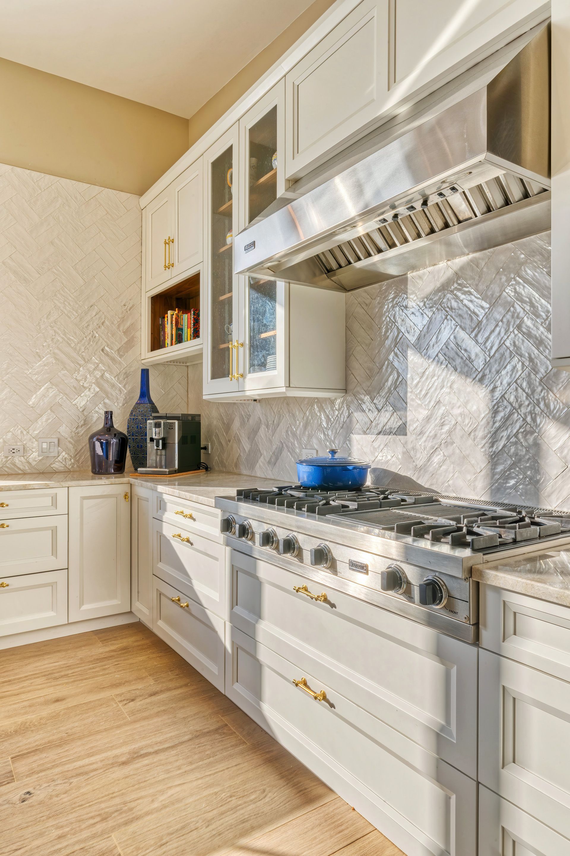 A bright kitchen with white cabinets, light countertops, and a stainless steel range. Herringbone backsplash and wood flooring add texture.