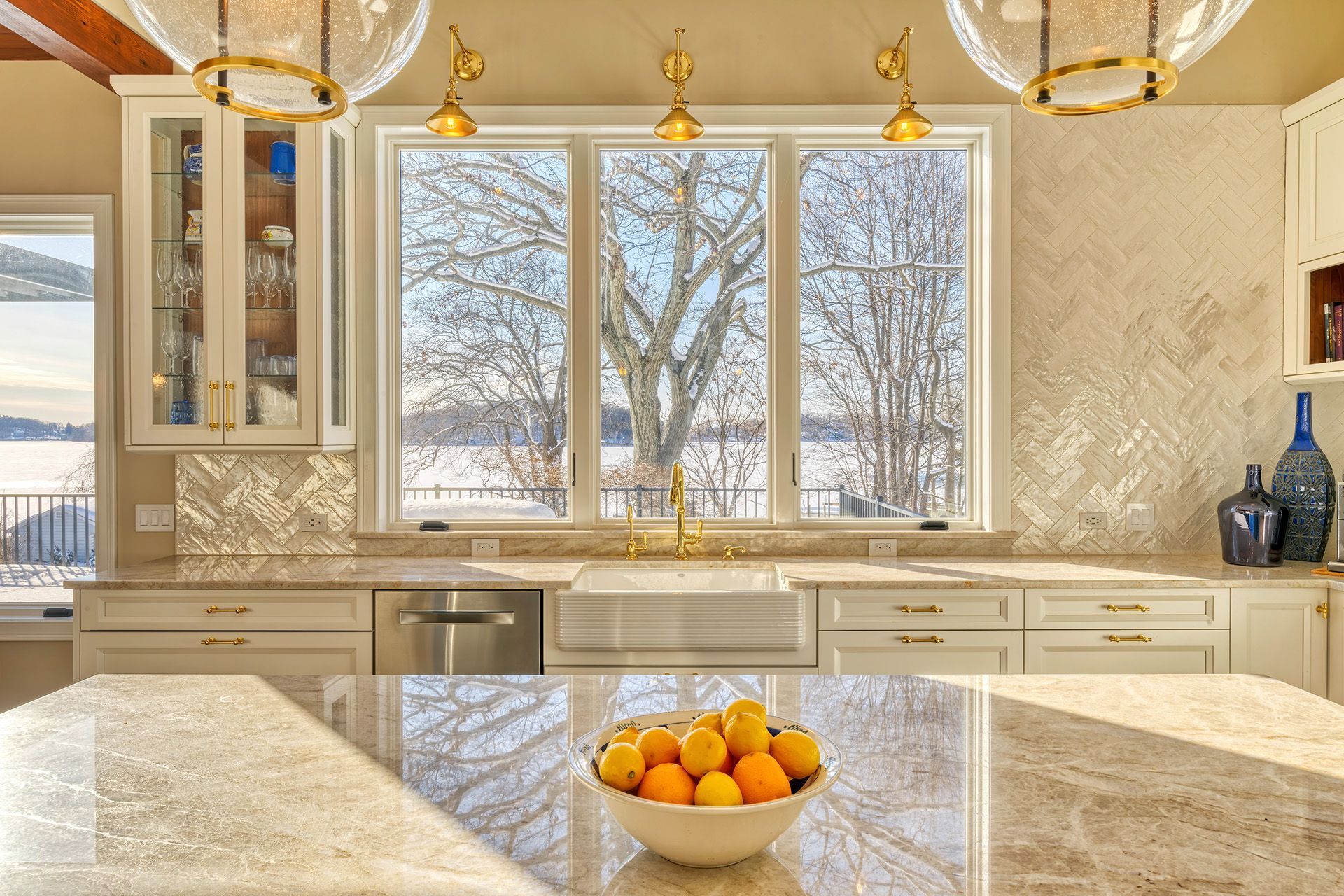 Kitchen with a sunny island, light countertops, and a large window overlooking a snowy landscape.  Gold fixtures and a bowl of lemons add warmth.