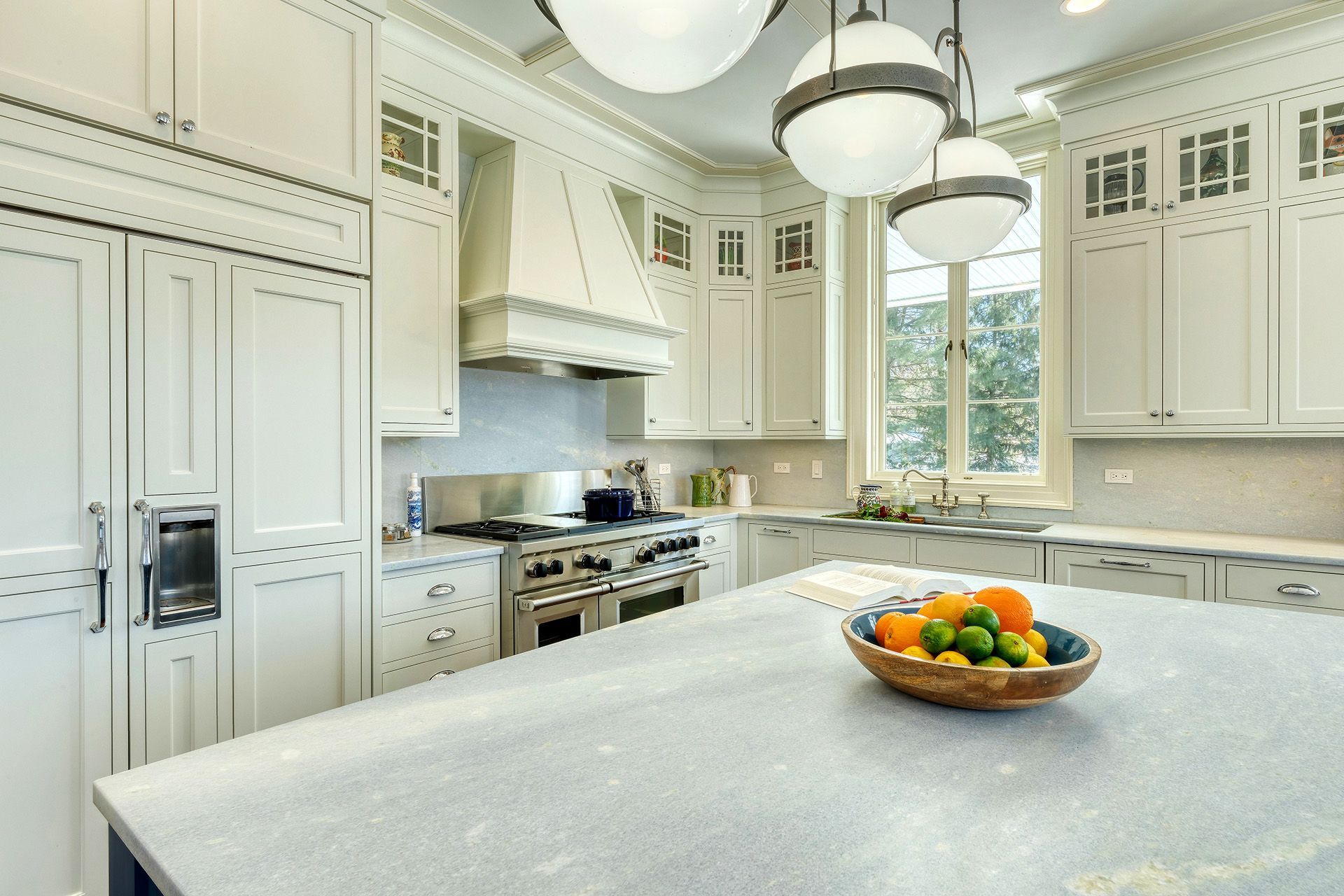 White kitchen with large island, stove, and overhead globe lights; a bowl of fruit rests on the island.