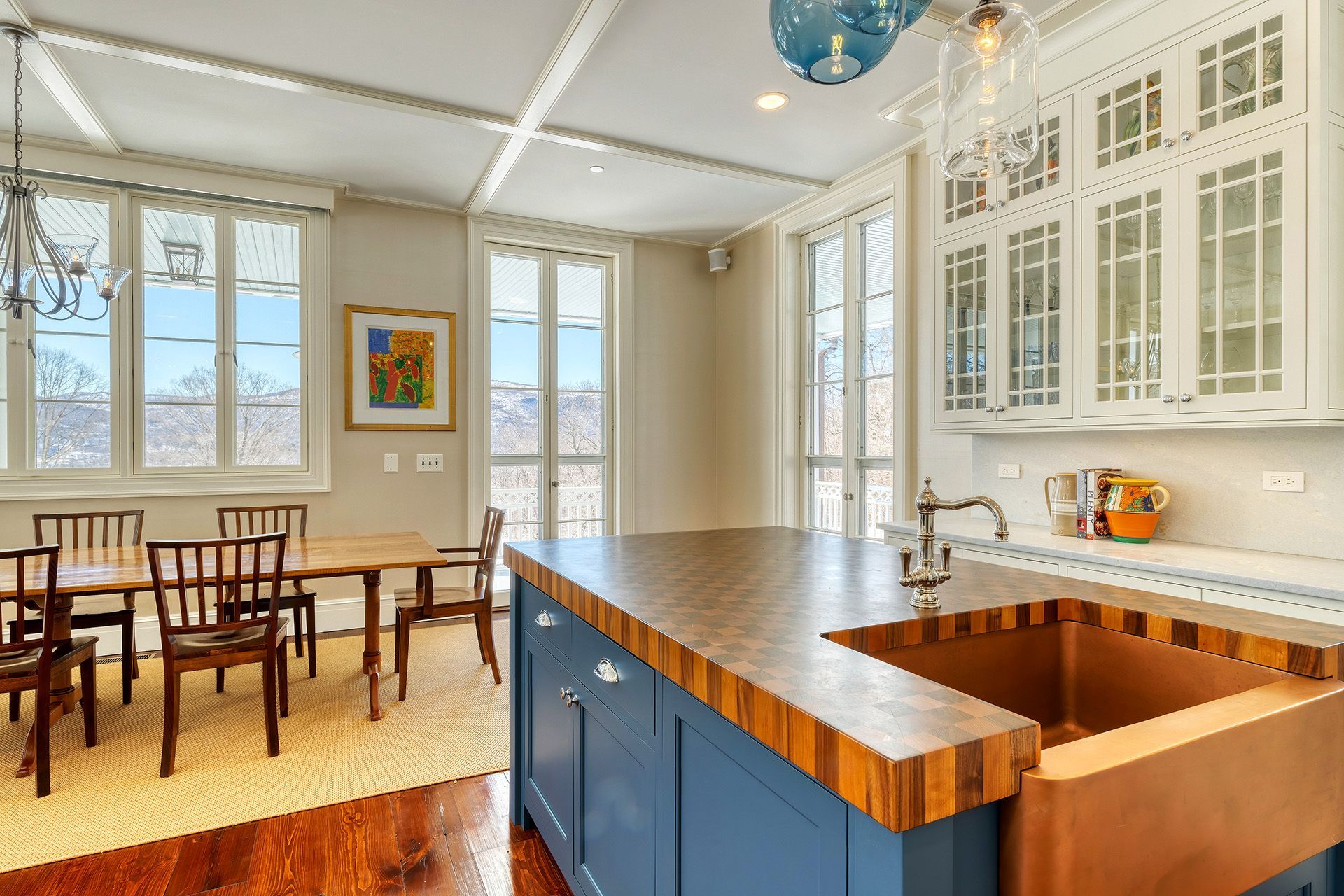 A bright, airy kitchen with a blue island, copper sink, and dining area. The room has large windows, white cabinets, and wooden accents.