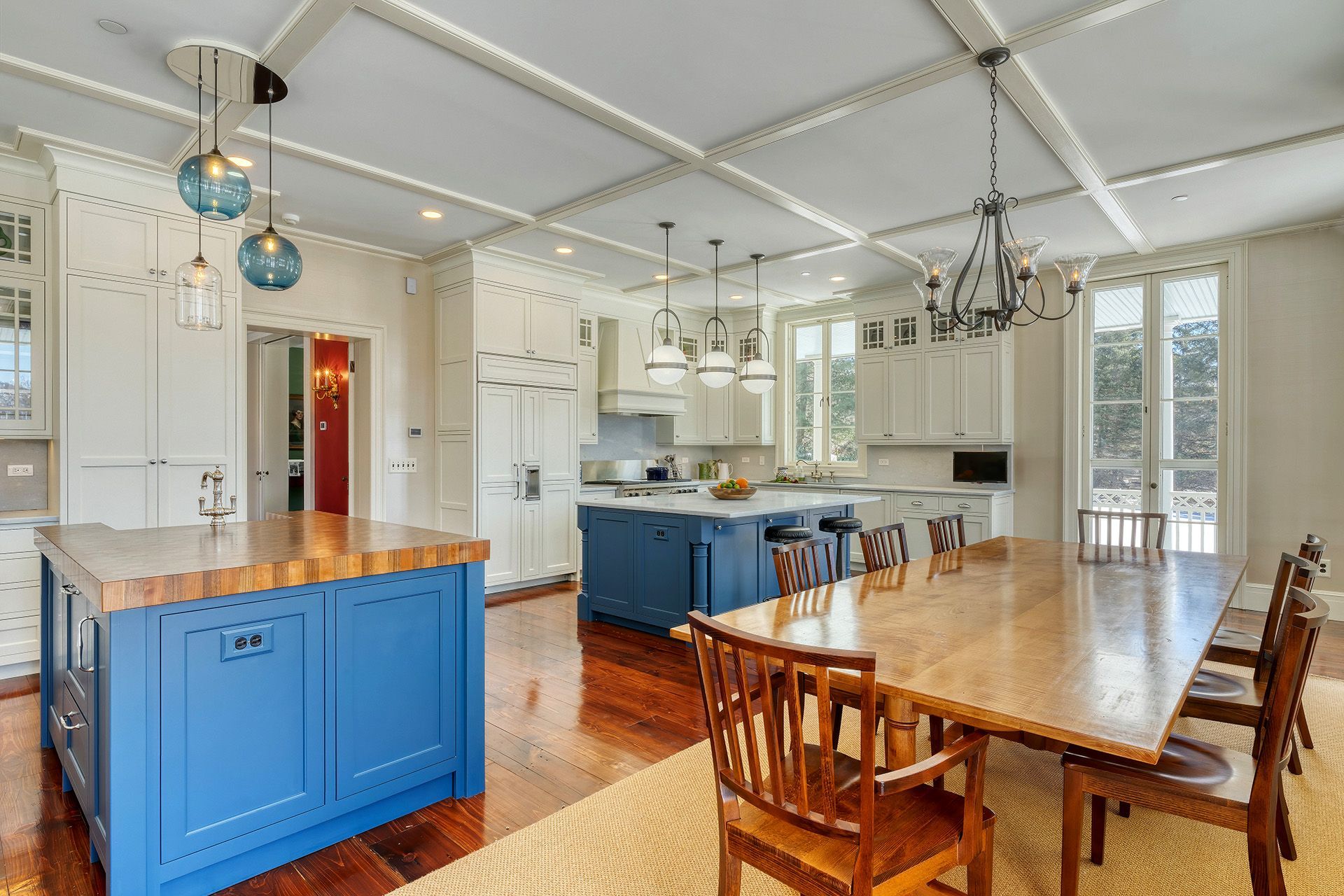 Spacious kitchen with blue islands, white cabinets, and a long wooden dining table. Light hardwood floors and a coffered ceiling are also visible.
