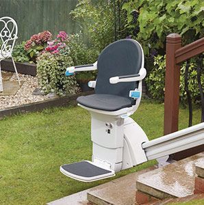 Stairlift on outdoor stairs; white chair with blue seat, armrests, and footrest. Brown railing, green grass, garden setting.