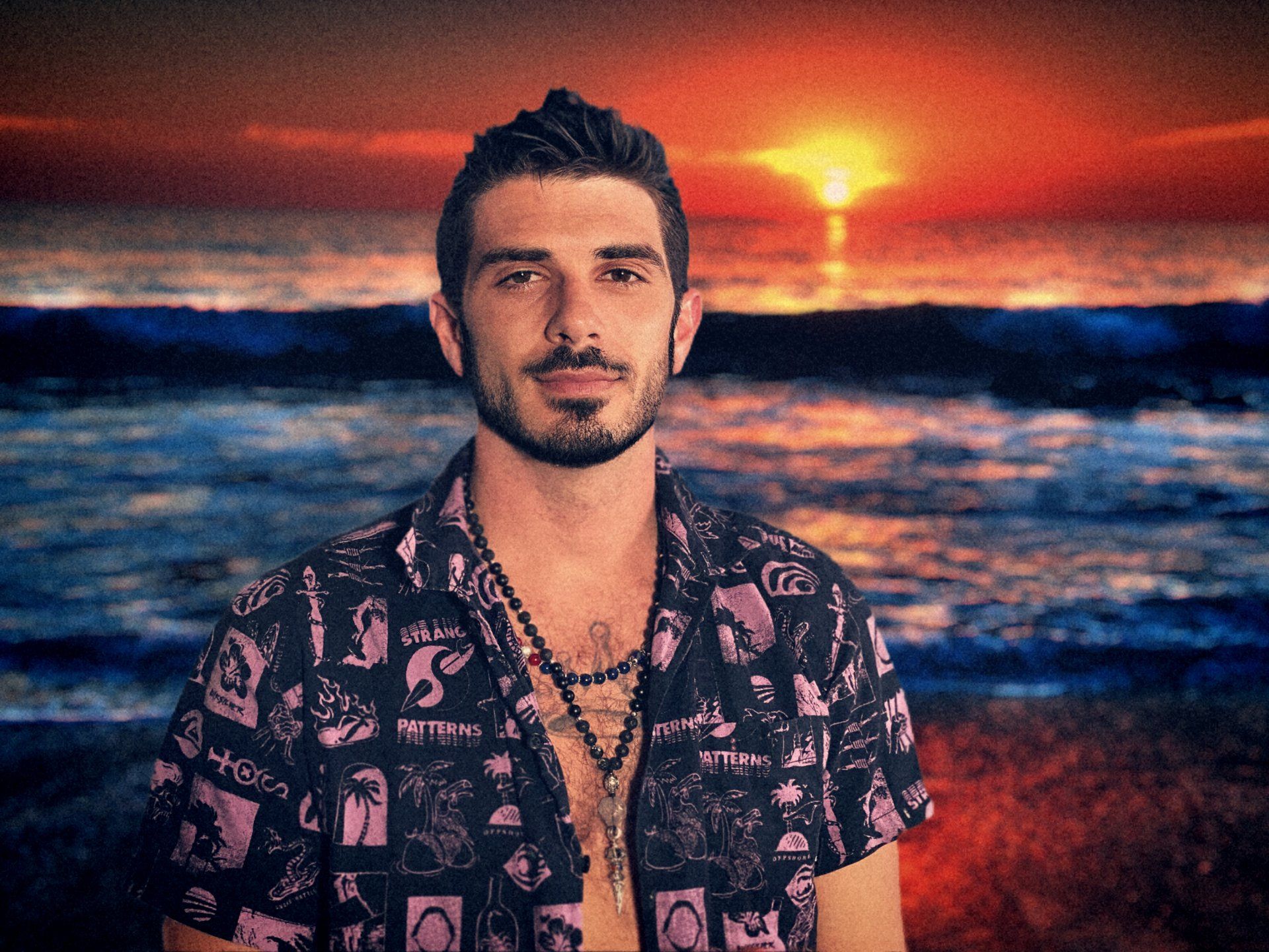 A man with a beard is standing on a beach at sunset.