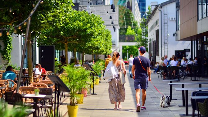 People walking along a sunny city street with outdoor café seating and potted plants.