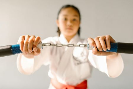 A young girl in a karate uniform is holding a pair of nunchaku.