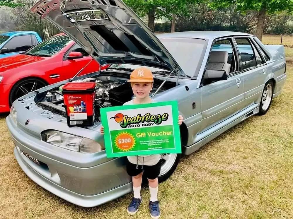 A Young Boy is Holding a Gift Voucher in Front of a Car — Seabreeze Auto Body in Coffs Harbour, NSW