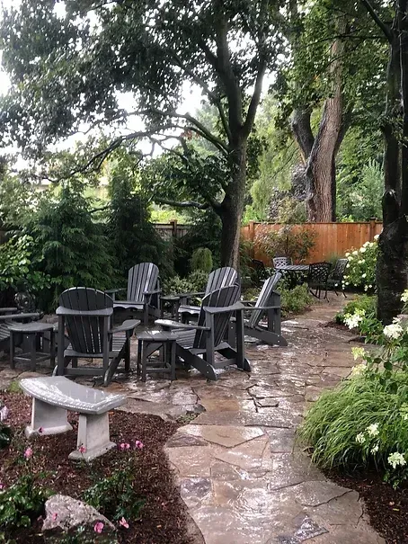 Outdoor patio with gray chairs on a stone path, surrounded by trees and flowers.