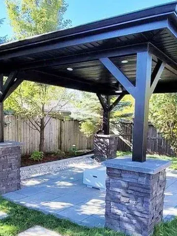 Black pergola with stone columns over a patio. Trees and a wooden fence are in the background.