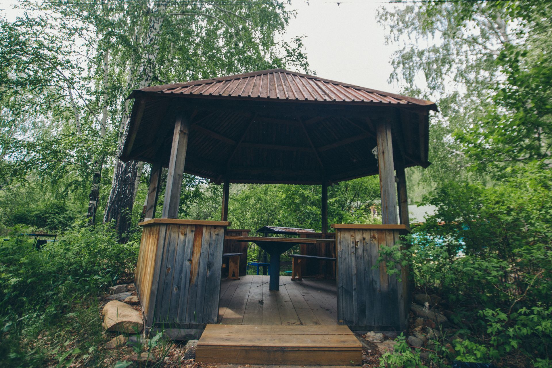 A wooden gazebo with a table and benches in the middle of a forest.