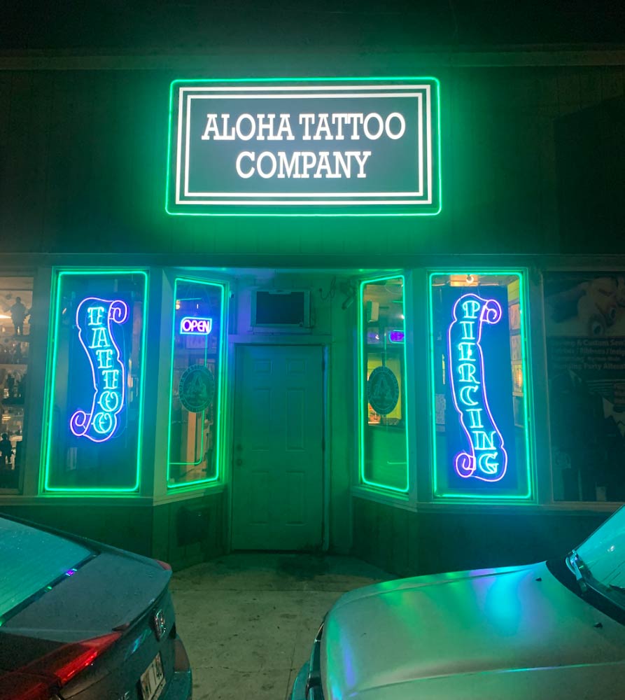 A neon sign for aloha tattoo company is lit up at night