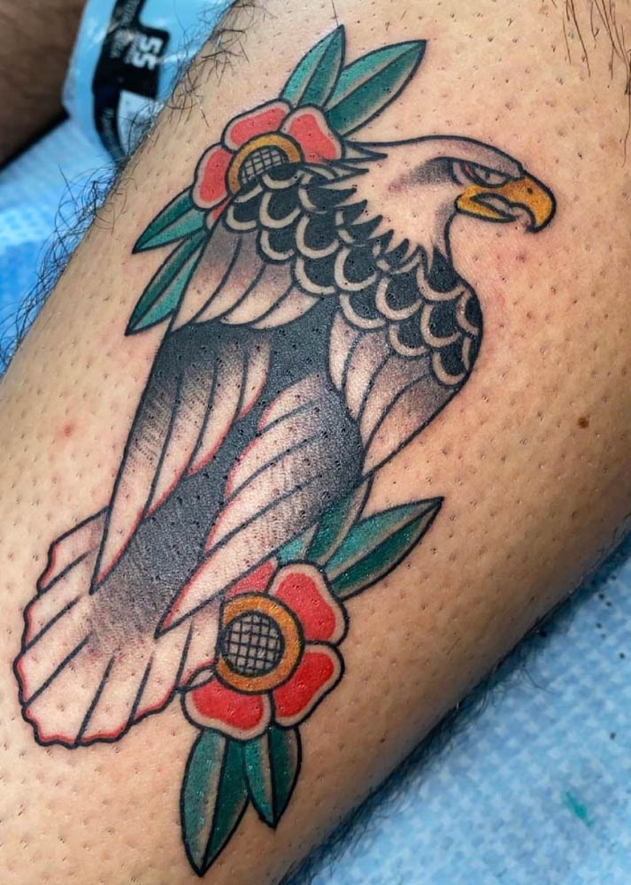 A tattoo of an eagle with flowers and leaves on a person 's leg.