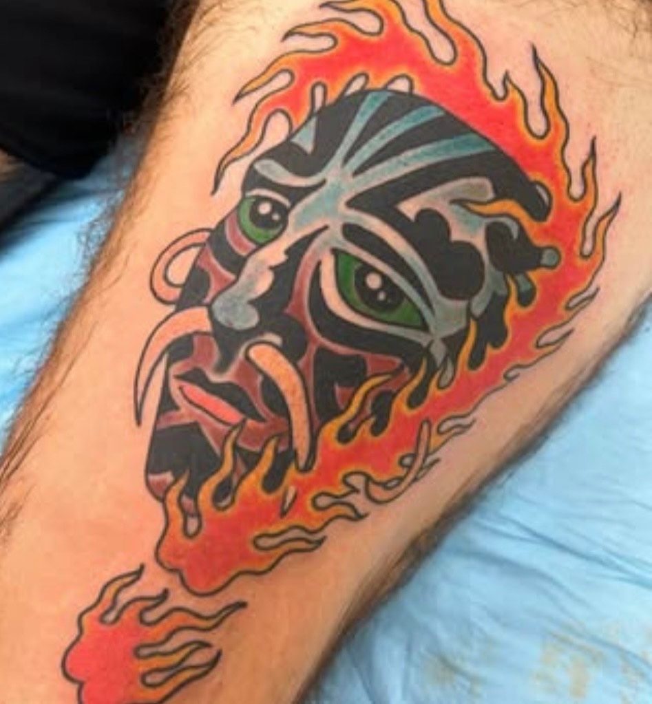 Tattoo by Aloha Tattoo of a stylized face with green eyes and fiery swirls on a calf.