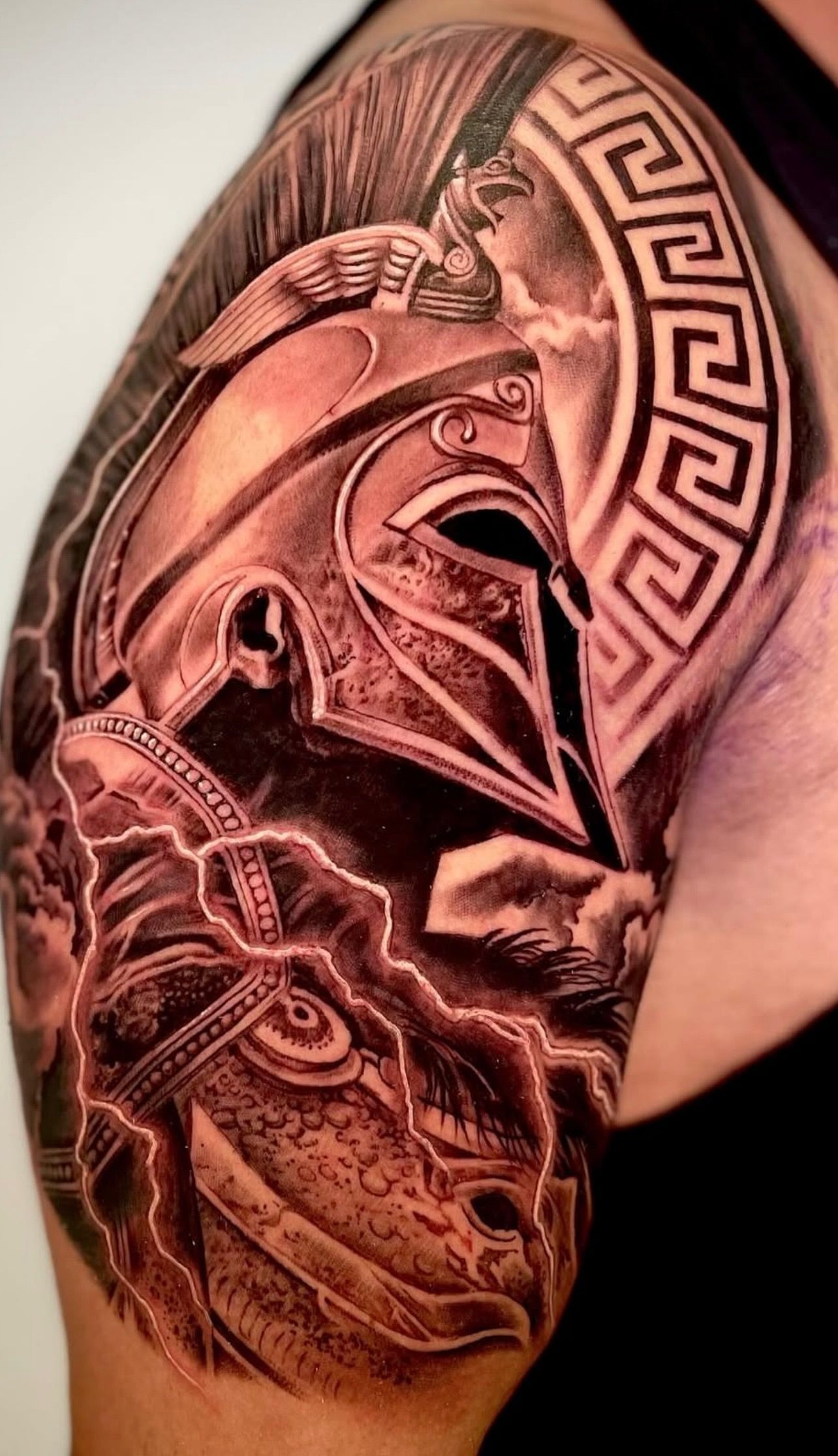 Upper arm tattoo of a Greek warrior helmet with winged accents, storm shading, and Greek key design—black and grey realism by guest artist Ricky Pantera.