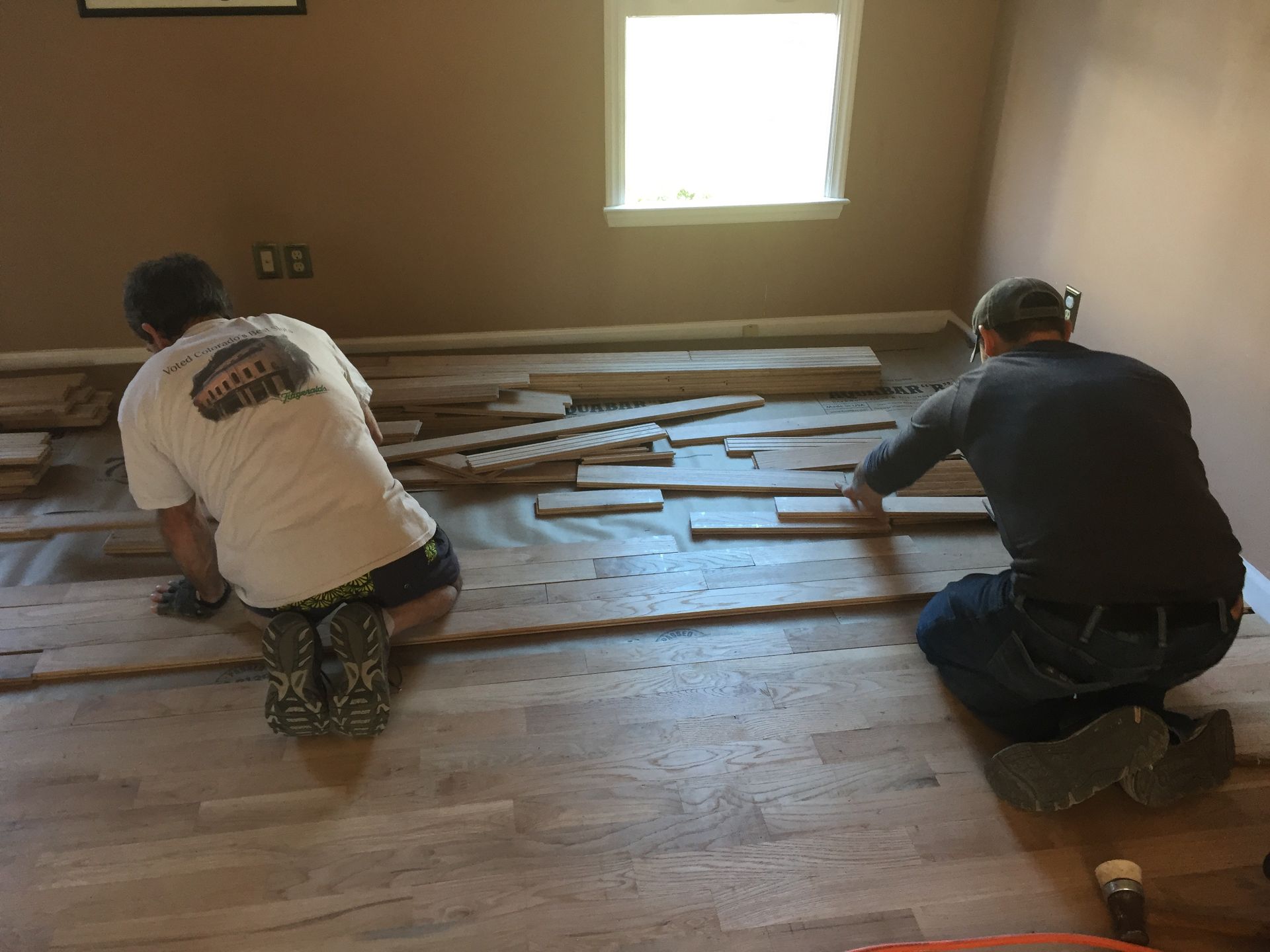 Two people installing hardwood flooring in a room with a window, kneeling and working.