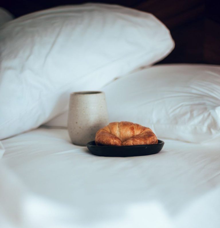 A croissant is on a plate on a bed next to a cup.