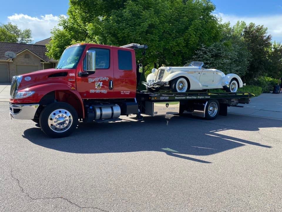 Red tow truck carrying a vintage white car on a sunny residential street.