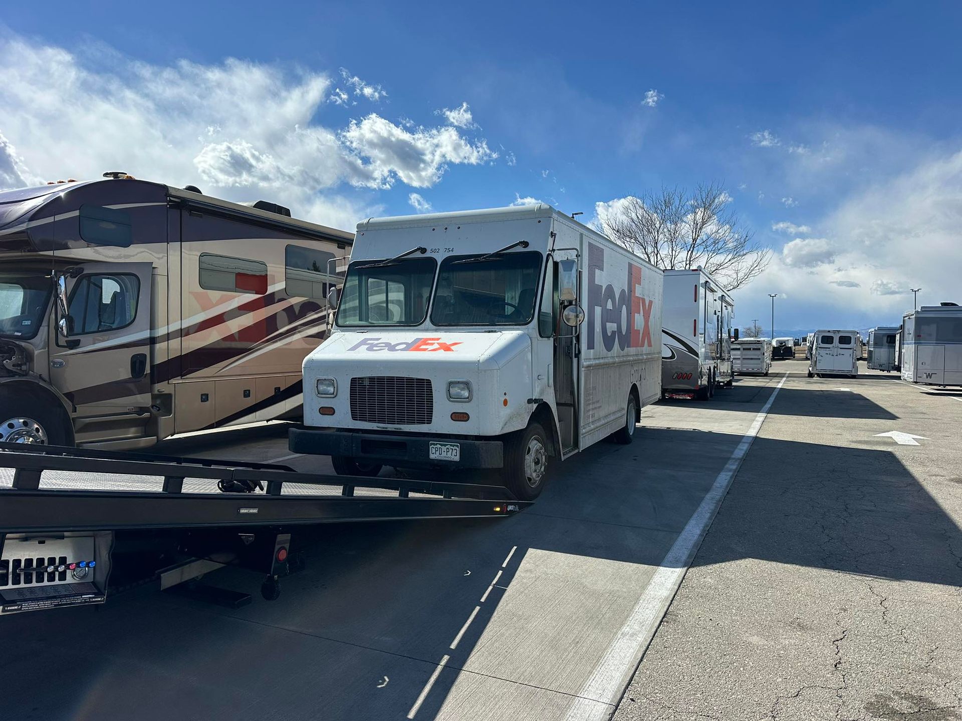 FedEx truck on a tow truck, parked on a paved lot, flanked by RVs and trailers under a blue sky.