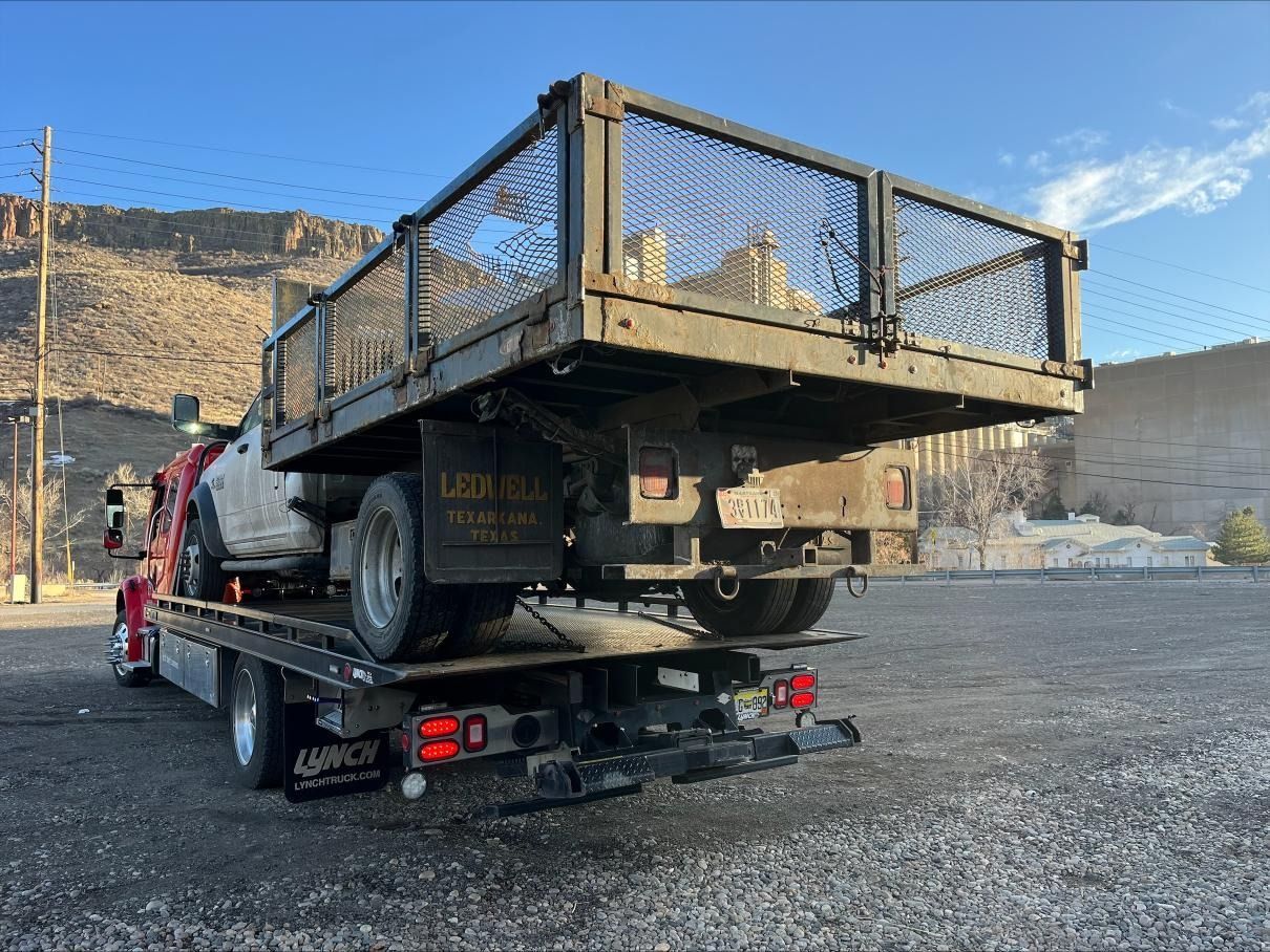 A flatbed tow truck carrying a white pickup truck on a gravel lot against a mountainous backdrop.