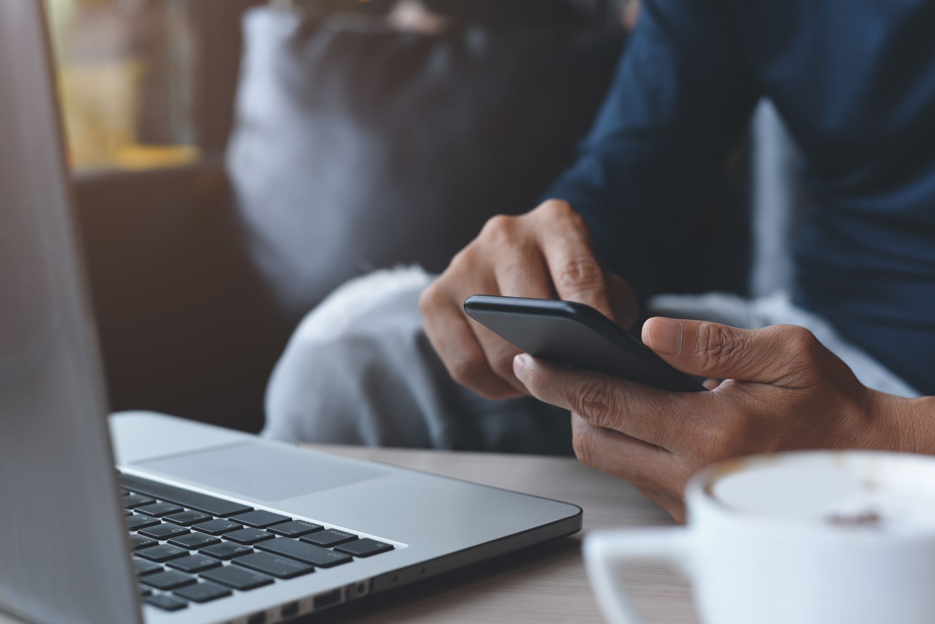 A person is sitting at a table using a cell phone and a laptop.
