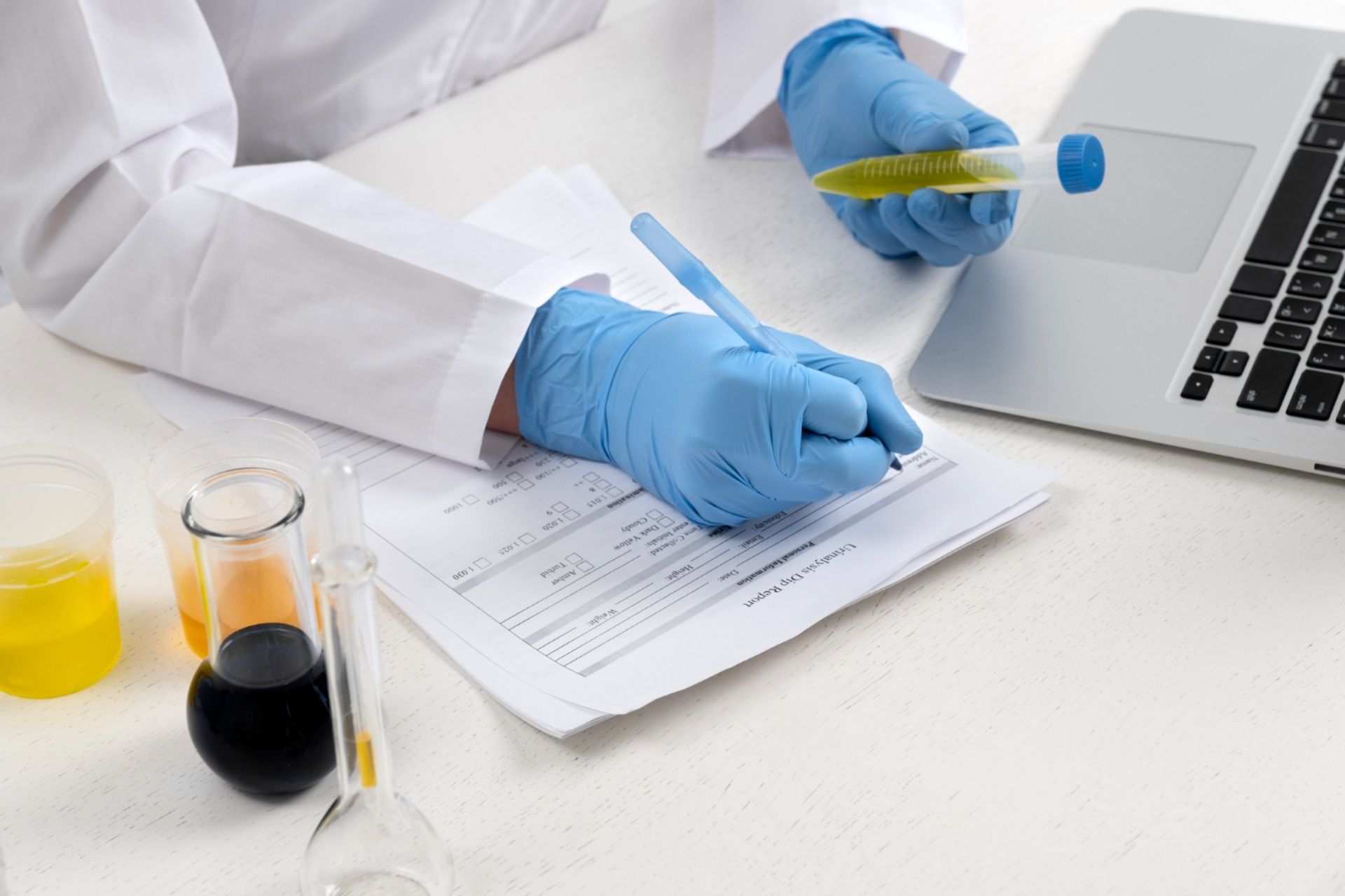 Scientist in lab coat and gloves taking notes while holding a test tube and using a laptop.