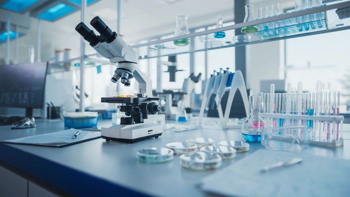 Microscope on a lab bench, surrounded by beakers, test tubes, and petri dishes; scientific lab environment.