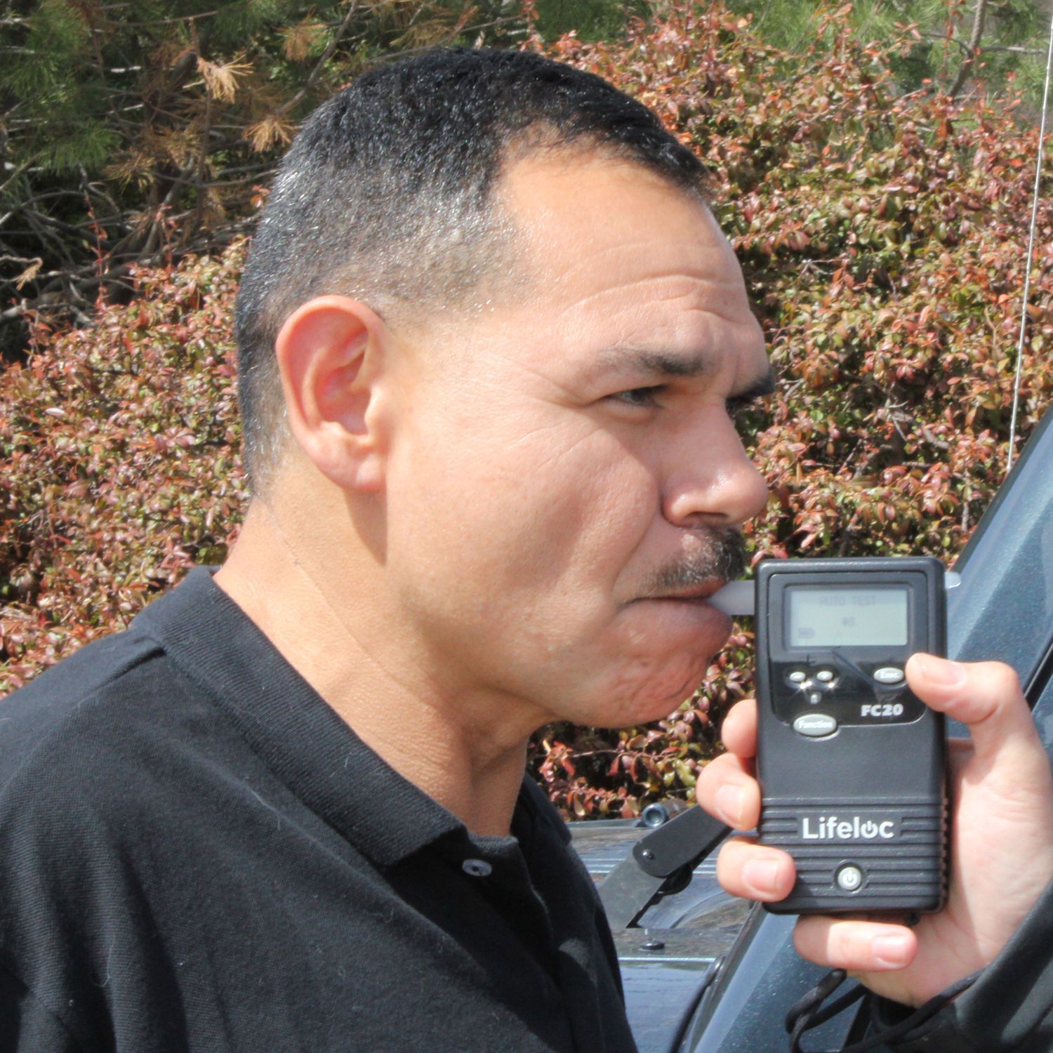 Man blowing into a breathalyzer test held by an arm outside. Outdoor setting.