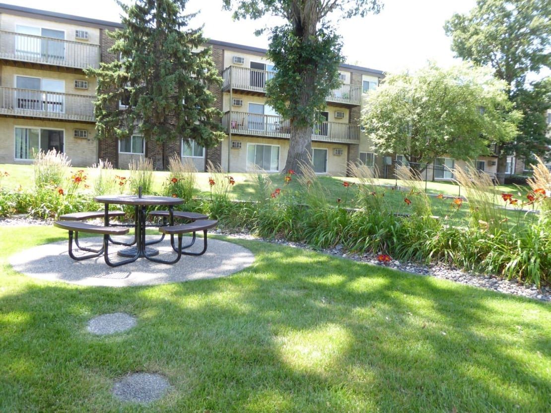 A picnic table is in the middle of a lush green field in front of a building.
