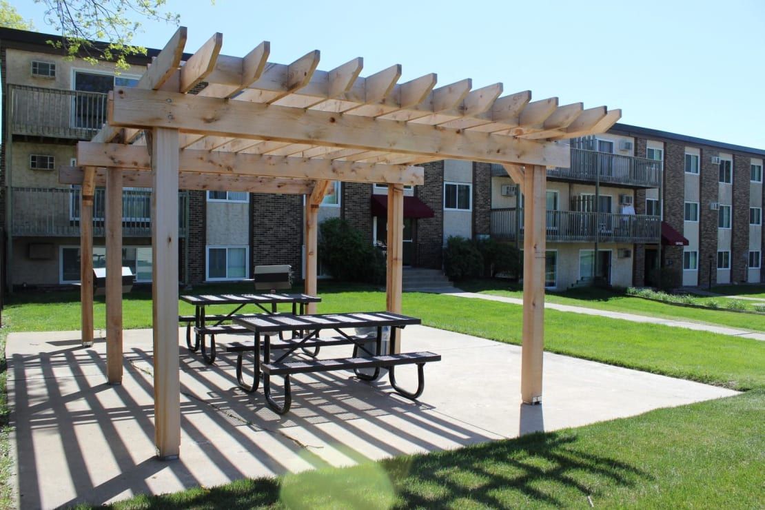 A wooden pergola with a picnic table underneath it in front of a building.